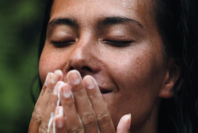 A close-up of a woman with wet skin and a radiant complexion, gently massaging a creamy cleanser onto her face. Her eyes are closed, and she has a relaxed, content expression. The background is blurred with hints of greenery, emphasising a natural and refreshing skincare moment.